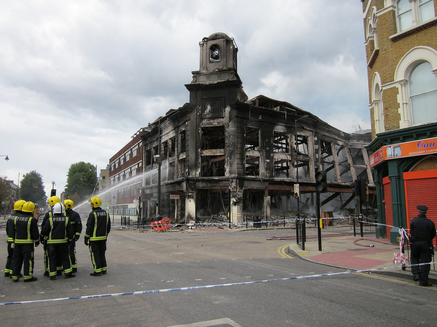 A gutted corner building on a London high street, its roof burned away and its upper floors reduced to a blackened frame. Five firefighters in helmets stand in the road playing a hose into the shell. Police tape crosses the foreground and an officer stands at the kerb on the right.